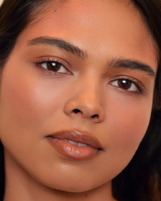 Model with deep skin wearing EM Cosmetics Pillow Plush Blush in Peanut for a soft nude flush.