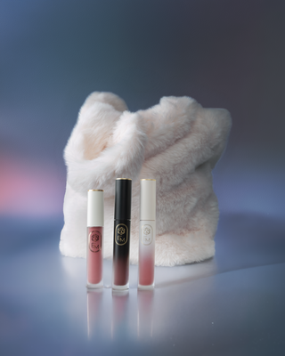 Still life editorial shot of three EM Cosmetics liquid lip products arranged on a textured surface.