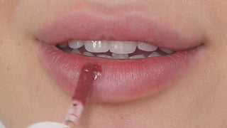 Model with dark skin wearing EM Cosmetics Everglass Lip Dew in Temptress for a glossy, deep berry lip look.