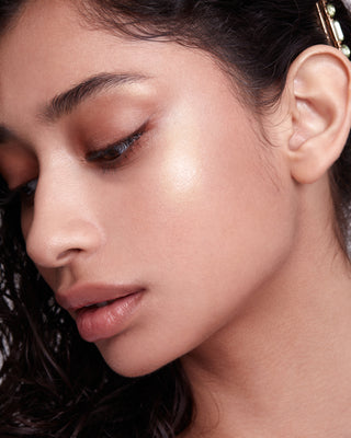 Model with medium-deep skin wearing EM Cosmetics Moonbeam Cushion Highlighter in Halo for a luminous gold shimmer.