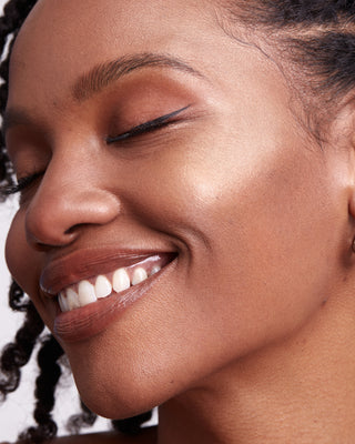 Model with dark skin wearing EM Cosmetics Moonbeam Cushion Highlighter in Divinity for a luminous high-point highlight.