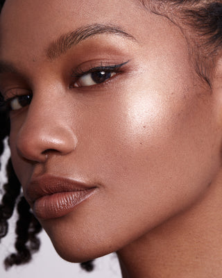 Model with dark skin wearing EM Cosmetics Moonbeam Cushion Highlighter in Halo for a rich, golden radiance.