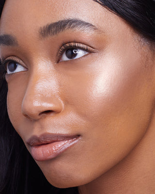 Model with dark skin wearing EM Cosmetics Sunscape Highlighter in Clarity for a high-contrast, luminous glow.