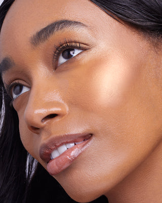 Model with dark skin wearing EM Cosmetics Sunscape Highlighter in Legendary for a luminous, golden cheek highlight.