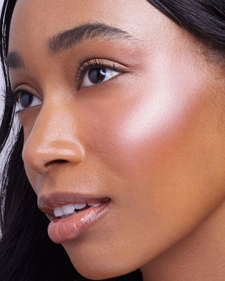 Model with dark skin wearing EM Cosmetics Sunscape Highlighter in Serenity for a luminous, rose gold glow.